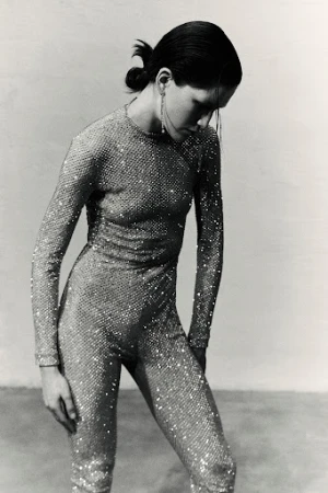 Monochrome fashion shot of a woman in a sparkling jumpsuit, long earrings, and a low ponytail against a neutral backdrop.