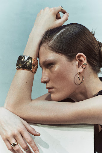 Close-up shot of an adult female model showcasing a gold cuff bracelet, gold hoop earrings, and rings in a bright, neutral setting.