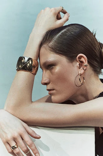 Close-up shot of an adult female model showcasing a gold cuff bracelet, gold hoop earrings, and rings in a bright, neutral setting.