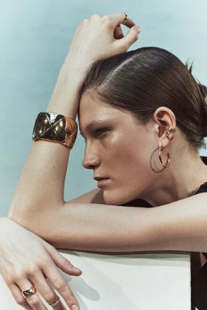 Close-up shot of an adult female model showcasing a gold cuff bracelet, gold hoop earrings, and rings in a bright, neutral setting.