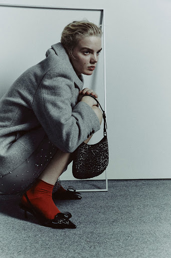 Model in a grey coat crouching with a black handbag, red socks, and black heels against a white seamless backdrop.