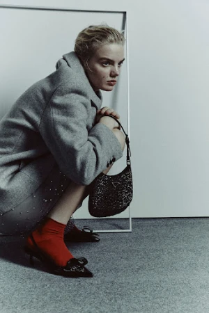 Model in a grey coat crouching with a black handbag, red socks, and black heels against a white seamless backdrop.