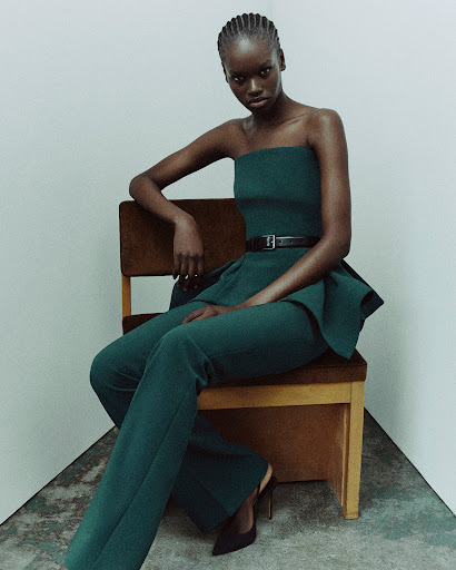 Fashion shot of a single African female model sitting on a chair wearing a teal dress and pants with a black belt against a plain white background.