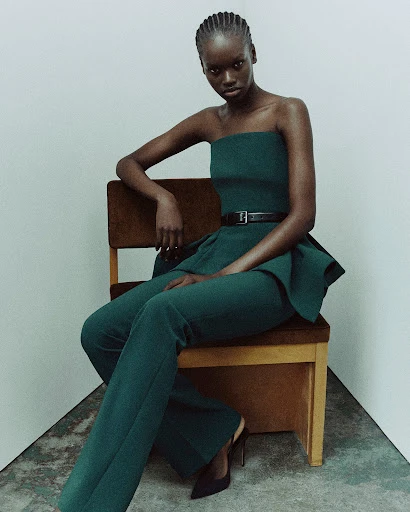 Fashion shot of a single African female model sitting on a chair wearing a teal dress and pants with a black belt against a plain white background.