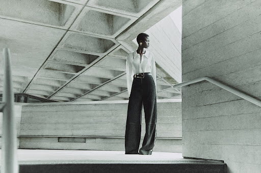 A slender African American woman in a white shirt and black pants stands on concrete steps against a brutalist architectural background.