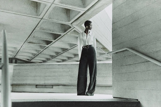 A slender African American woman in a white shirt and black pants stands on concrete steps against a brutalist architectural background.
