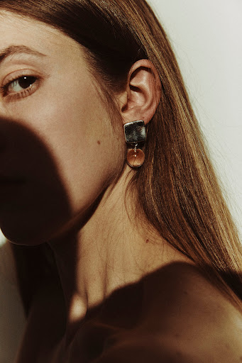 Close-up portrait of a woman with auburn hair wearing modern earrings, featuring geometric shapes and a translucent bead detail.