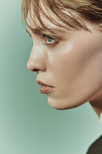 Close-up portrait of a young adult woman with fair skin, light brown hair, and blue eyes against a light green background.