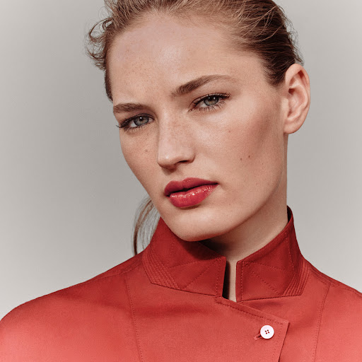 Close-up portrait of a woman in her 20s wearing a red high-collared jacket against a neutral grey seamless background