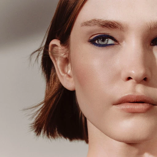 Close-up portrait of a fair-skinned woman with auburn hair and blue eyeliner makeup against a neutral gray studio background.