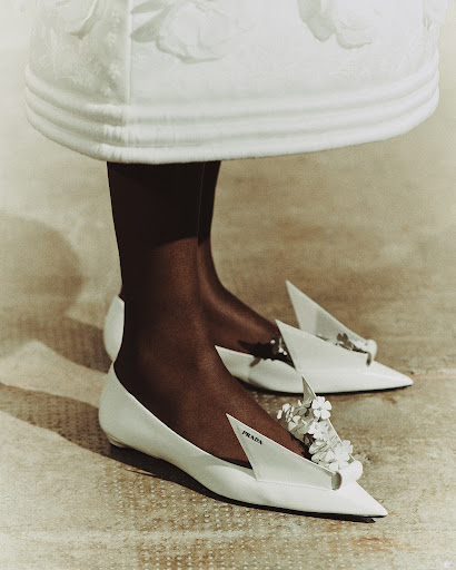 Close-up of a pair of white Prada shoes with floral embellishments worn by a black woman, complemented by a white dress.