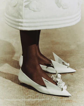 Close-up of a pair of white Prada shoes with floral embellishments worn by a black woman, complemented by a white dress.