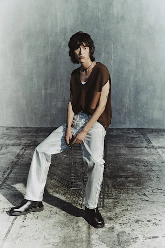 A young adult male sits indoors on a metal grid seat wearing a brown short-sleeve knit shirt, light blue jeans, and black leather boots.