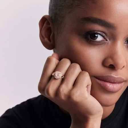 Close-up of an adult woman of color wearing a rose-gold engagement ring with diamond halo, posing in front of a neutral grey backdrop.
