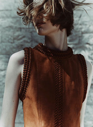 Indoor shot of a stylish brunette woman wearing a brown suede vest with braided trim, posing against a textured gray-blue wall in natural light.