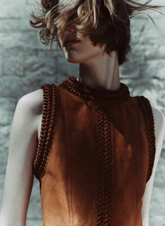 Indoor shot of a stylish brunette woman wearing a brown suede vest with braided trim, posing against a textured gray-blue wall in natural light.