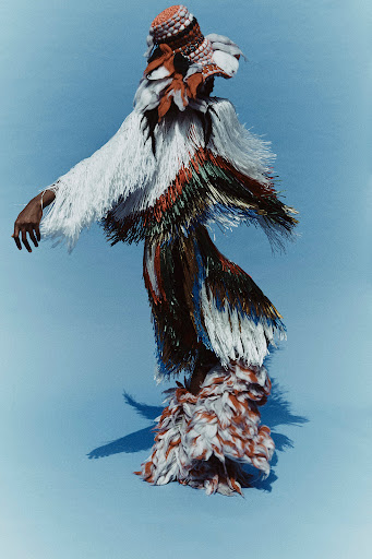 Fashionable person wearing a unique fringed garment with colorful accents, complemented by a textured hat, posing on blue background.