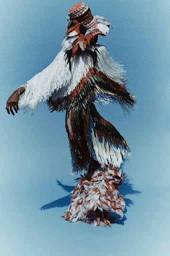 Fashionable person wearing a unique fringed garment with colorful accents, complemented by a textured hat, posing on blue background.