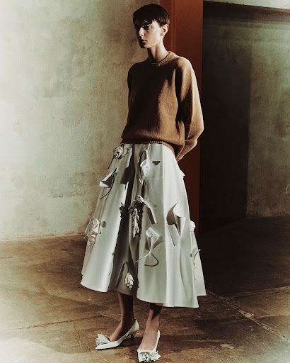 An adult female model with short hair wears a brown sweater and white skirt, standing in a gritty interior space for a fashion shot.