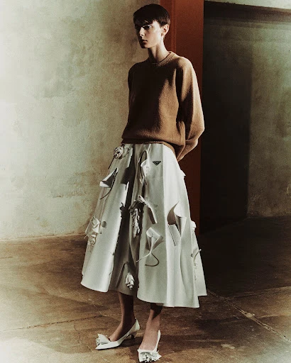 An adult female model with short hair wears a brown sweater and white skirt, standing in a gritty interior space for a fashion shot.