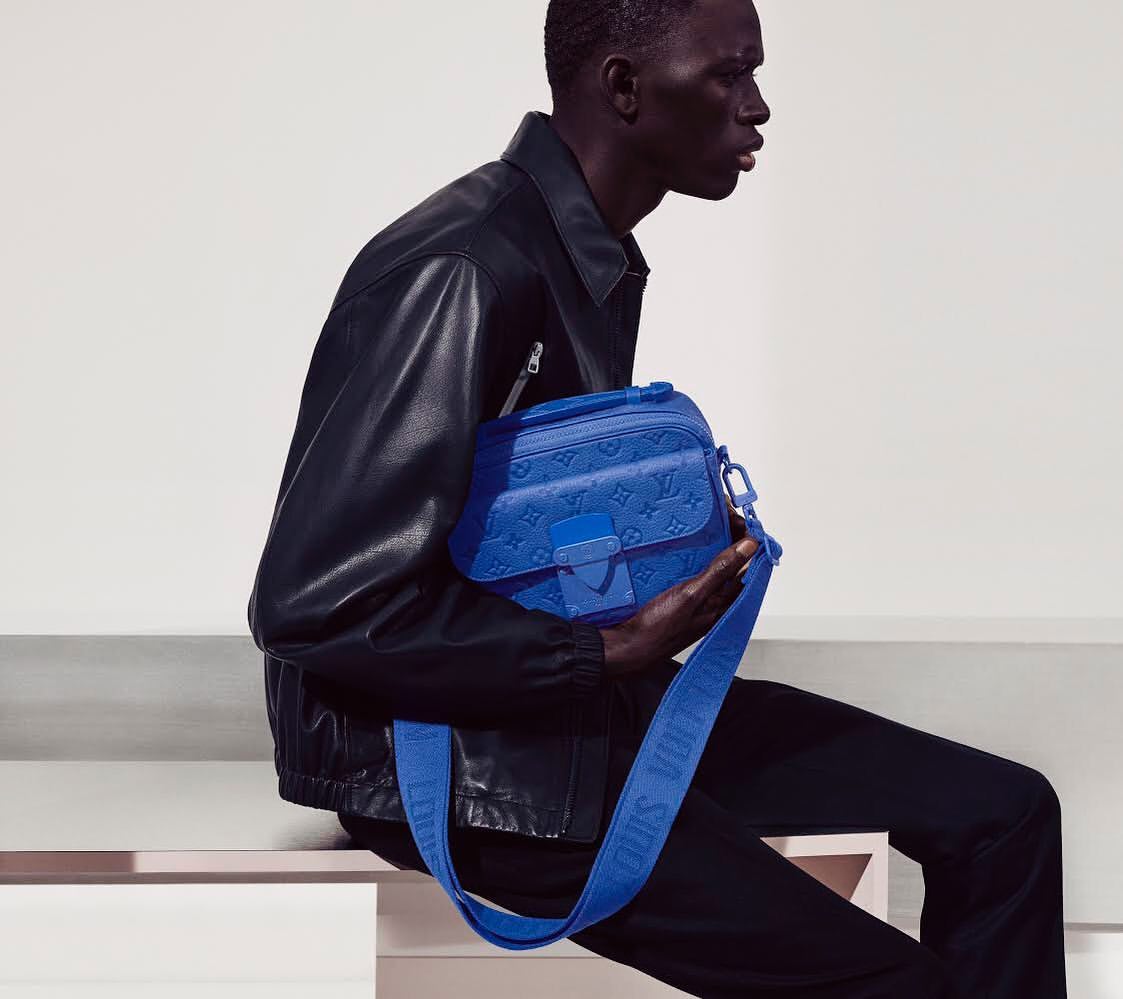 Stylish black male model sitting and holding blue Louis Vuitton bag in a studio shot with a white background and fashionable styling.