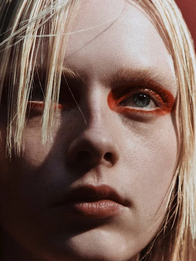 Close-up portrait of a blonde female model with light skin and orange makeup accentuating her eyes, against a red backdrop.