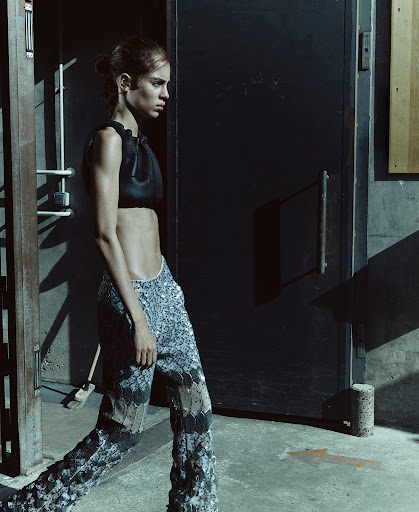 A woman in a black top and patterned pants walks past a metal door in an industrial setting, showcasing fashion and style.