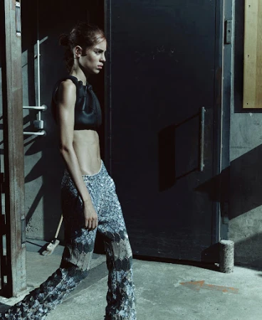 A woman in a black top and patterned pants walks past a metal door in an industrial setting, showcasing fashion and style.