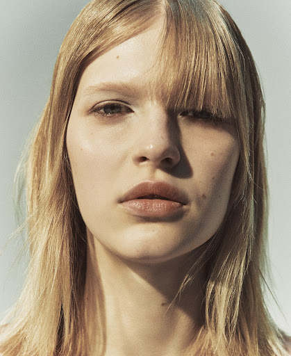 Close-up studio portrait of a young adult woman with blonde hair, wearing neutral makeup and looking directly at the camera against a light backdrop.