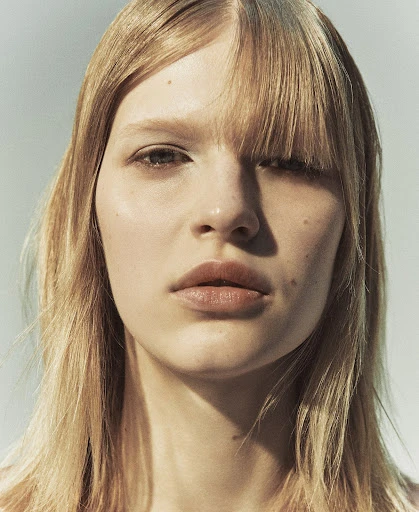 Close-up studio portrait of a young adult woman with blonde hair, wearing neutral makeup and looking directly at the camera against a light backdrop.