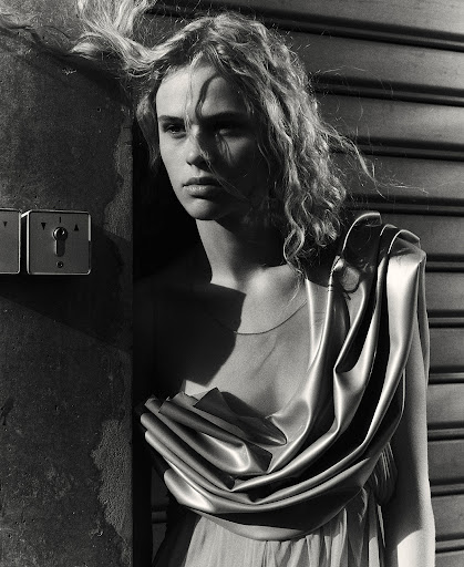 Monochrome portrait of a young woman with wavy hair in a draped gown, leaning against a textured wall with industrial switch plates.