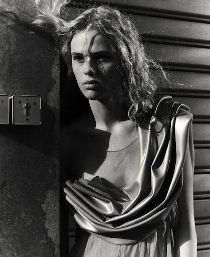Monochrome portrait of a young woman with wavy hair in a draped gown, leaning against a textured wall with industrial switch plates.