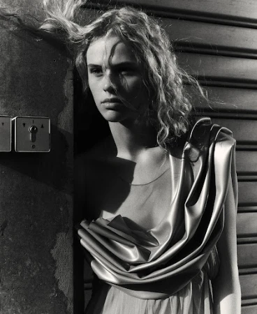 Monochrome portrait of a young woman with wavy hair in a draped gown, leaning against a textured wall with industrial switch plates.