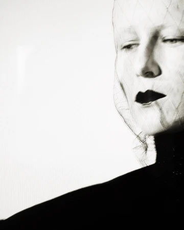 Black and white artistic portrait of a fair-skinned woman with dark lipstick and a net-like structure over her face.