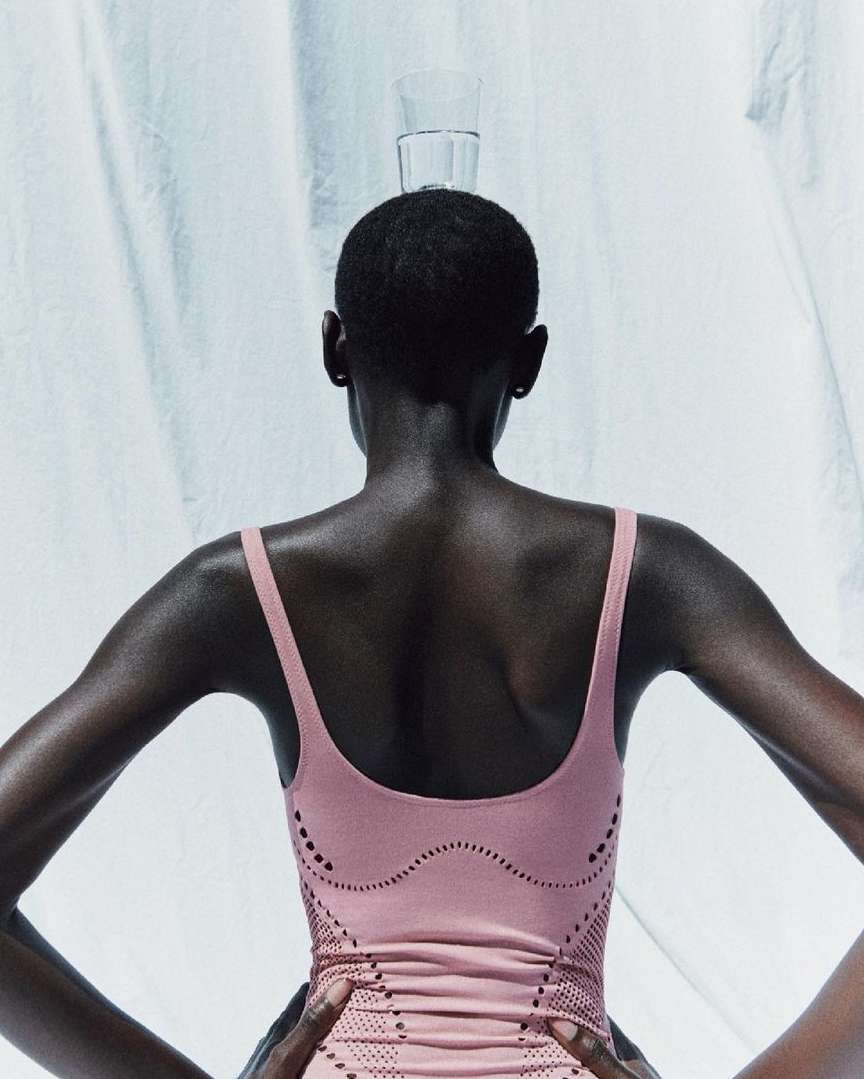 Back view of a dark-skinned woman in pink fitness wear balancing a glass of water on her head against a light backdrop.