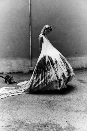 Black and white editorial image of a woman in a long white dress standing outdoors with a cat nearby.
