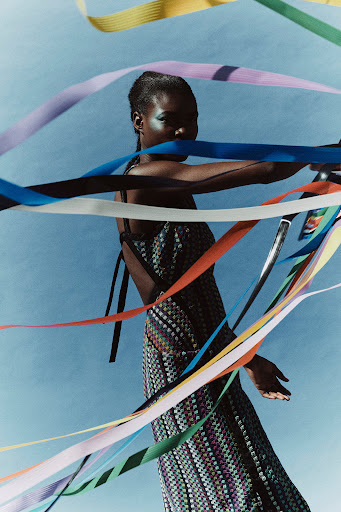 A full shot of an adult woman in a patterned dress, surrounded by colorful ribbons, posing in front of a light blue background in a studio.