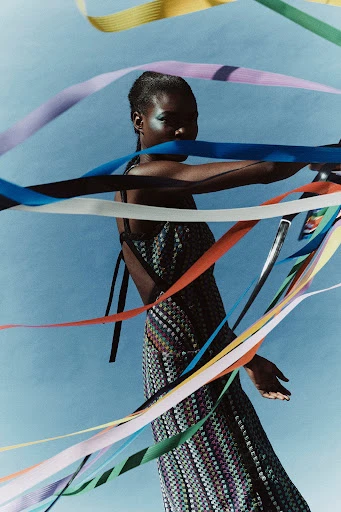 A full shot of an adult woman in a patterned dress, surrounded by colorful ribbons, posing in front of a light blue background in a studio.