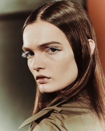 Close-up beauty portrait of a woman with long straight brown hair, blue feathered eyelashes, and wearing an olive green jacket.