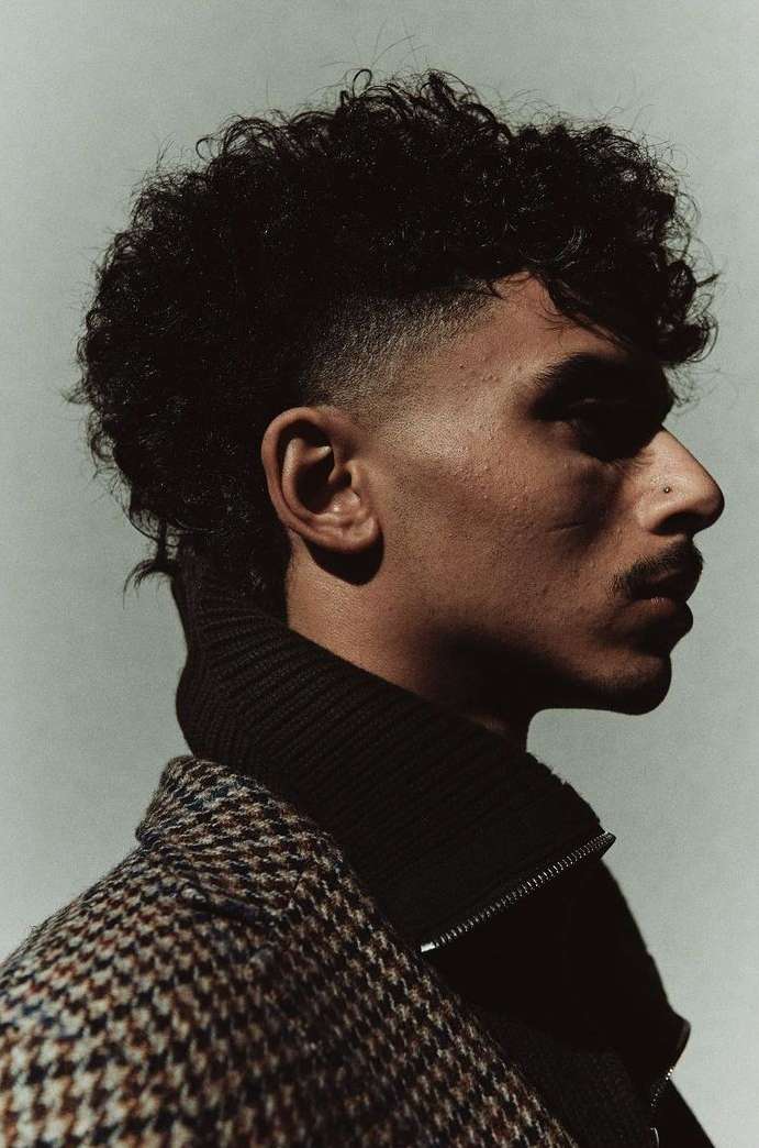 Profile portrait of a man with curly hair wearing a houndstooth jacket and a black turtleneck against a light background in a photo studio.
