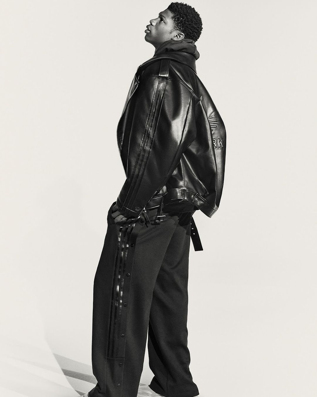 A young Black male model poses indoors wearing a black leather jacket and black side-button track pants in a fashion studio shoot.