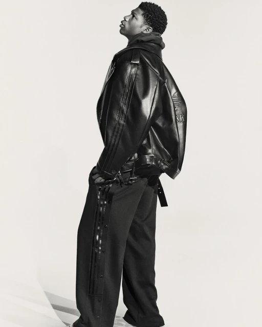 A young Black male model poses indoors wearing a black leather jacket and black side-button track pants in a fashion studio shoot.