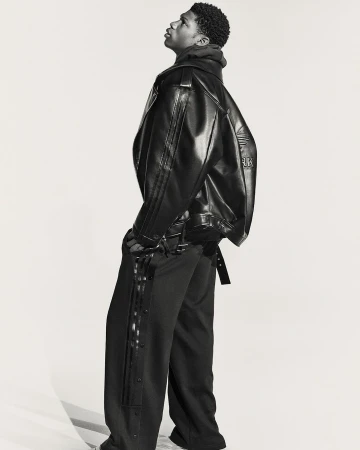 A young Black male model poses indoors wearing a black leather jacket and black side-button track pants in a fashion studio shoot.