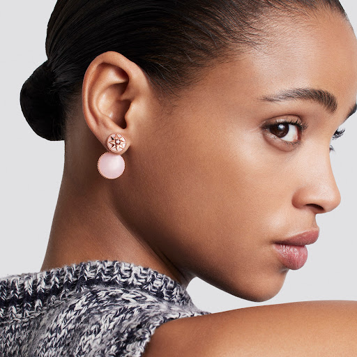 Close-up portrait of a woman wearing pink drop earrings and a grey textured knit top against a neutral light grey background