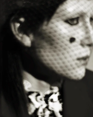Close-up portrait of a woman with a veil featuring polka dots, wearing a dark jacket with a patterned collar. Blurred effect.