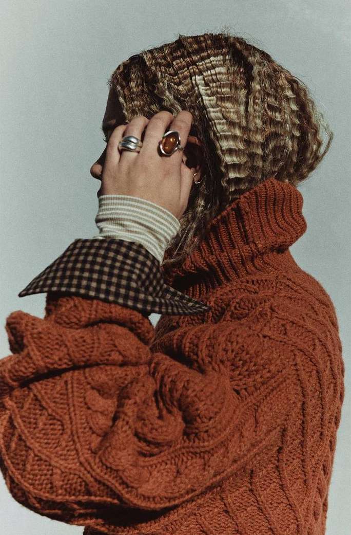 Profile shot of a woman with crimped blonde hair, wearing an orange cable knit sweater and silver rings against a white background.
