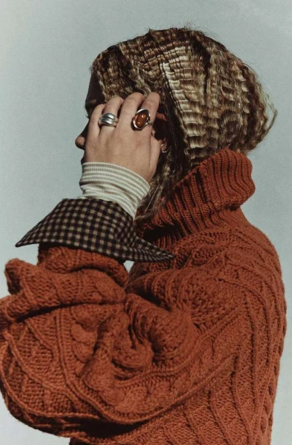 Profile shot of a woman with crimped blonde hair, wearing an orange cable knit sweater and silver rings against a white background.