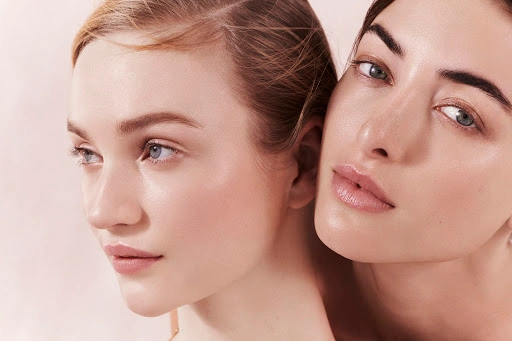 Close-up portrait of two female models with radiant skin and soft natural makeup posed close together against a pale background