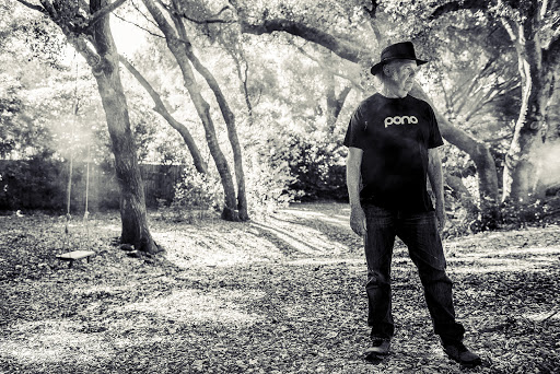 Black and white portrait of a senior man standing outside in a wooded area with trees, wearing a hat and T-shirt with jeans.