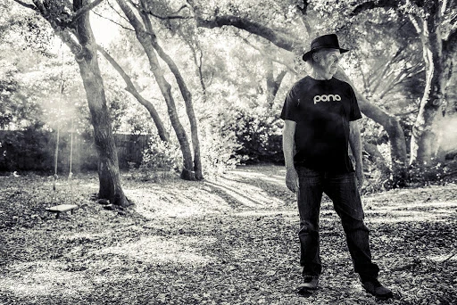 Black and white portrait of a senior man standing outside in a wooded area with trees, wearing a hat and T-shirt with jeans.